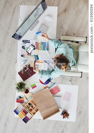 High angle view of young woman in hoodie sitting at desk and viewing palettes while picking out colors for new dresses 91990894