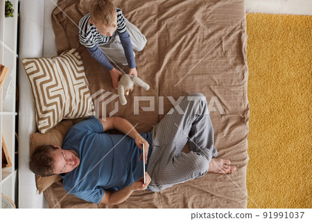High angle view of relaxed young father in pajamas watching video on tablet while his son playing toy, they spending time in bed 91991037