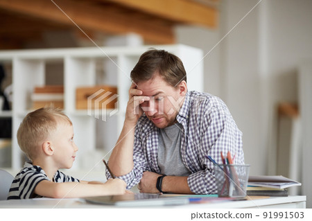 Confused young father puzzled with sons home task solution sitting at table and holding head in hand 91991503