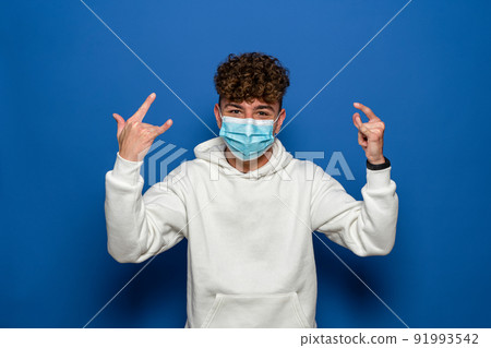 Young man with curly hair wearing a face mask for coronavirus making the rock gesture isolated on blue studio background 91993542