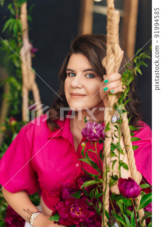 a brunette woman in a pink blouse with peonies on the swing. a brunette woman in a pink blouse with peonies on the swing. 91994585