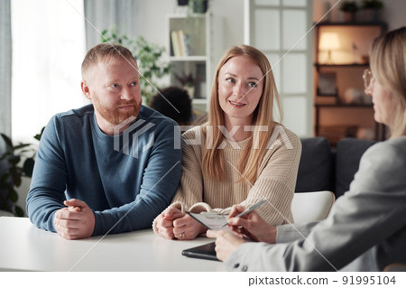Young couple having an interview with social worker before adoption of child at table during meeting while she making notes in document Young couple having an interview with social worker before adoption of child at table during meeting while she making notes in document 91995104