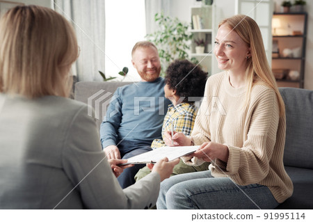 Happy young woman signing contract about adoption while talking to social worker with her family in background Happy young woman signing contract about adoption while talking to social worker with her family in background 91995114