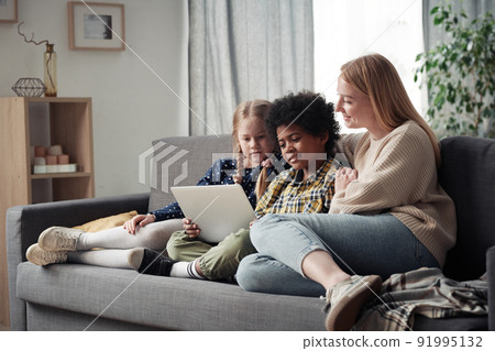 Young mother resting on sofa with her two adoptive children and they watching movie together using laptop Young mother resting on sofa with her two adoptive children and they watching movie together using laptop 91995132