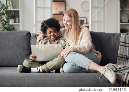 Young mother watching cartoon on laptop with her little adoptive son while they resting on sofa in living room Young mother watching cartoon on laptop with her little adoptive son while they resting on sofa in living room 91995133