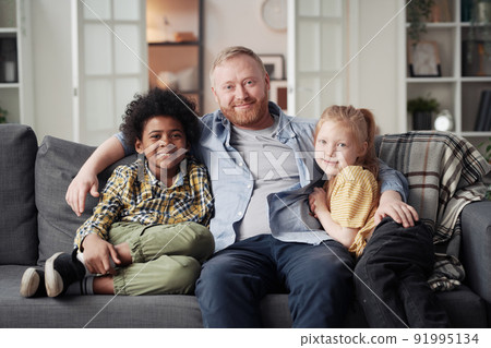 Portrait of young father embracing his adopted children while they resting on sofa in living room Portrait of young father embracing his adopted children while they resting on sofa in living room 91995134