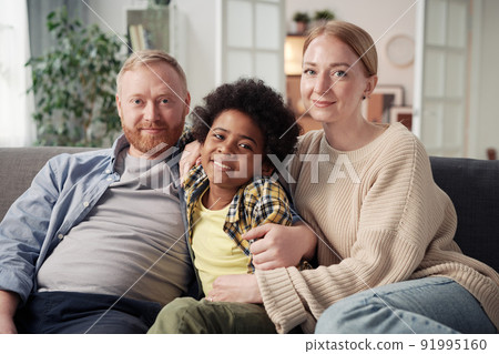 Portrait of young parents embracing their adopted son African American ethnicity and smiling at camera while sitting on sofa Portrait of young parents embracing their adopted son African American ethnicity and smiling at camera while sitting on sofa 91995160