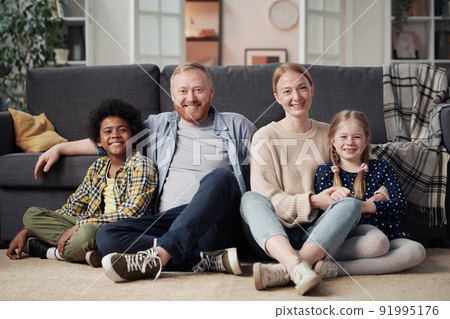 Portrait of happy family with adopted children sitting on floor in living room and smiling at camera Portrait of happy family with adopted children sitting on floor in living room and smiling at camera 91995176