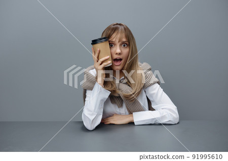 Shocked happy young blonde student lady freelancer in warm sweater with takeaway coffee sitting open mouth in gray modern home office. Coffee break Lover Concept. Copy space. Cool offer Shocked happy young blonde student lady freelancer in warm sweater with takeaway coffee sitting open mouth in gray modern home office. Coffee break Lover Concept. Copy space. Cool offer 91995610
