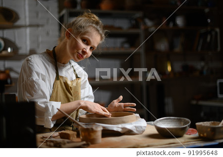 A ceramist makes a plate. Woman in an apron works in a pottery workshop. A ceramist makes a plate. Woman in an apron works in a pottery workshop. 91995648