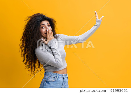 LOOK HERE. Surprised confused young arab lady covers her mouth with hand, points finger up, shows copy space, isolated on yellow background. Shocking offer, news, great sale, advertising and app LOOK HERE. Surprised confused young arab lady covers her mouth with hand, points finger up, shows copy space, isolated on yellow background. Shocking offer, news, great sale, advertising and app 91995661