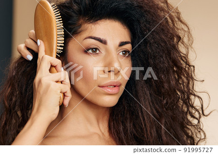 HEALTHY LONG HAIR TREATMENT CONCEPT. Pensive happy adorable tanned awesome curly Latin lady with hairbrush combing curls posing isolated on pastel beige background, look aside. Copy space, good offer 91995727