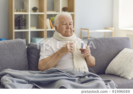 Sick senior man with a glass of water in his hand suffers from a severe cold sitting on the couch. Sick senior man with a glass of water in his hand suffers from a severe cold sitting on the couch. 91996089