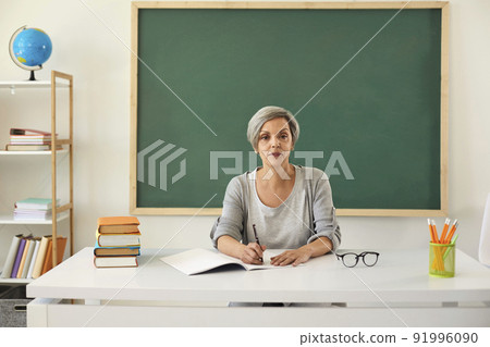 Teacher teaches. Serious senior woman teacher looking at the camera at lesson on the background of a greenboard in the classroom. Teacher teaches. Serious senior woman teacher looking at the camera at lesson on the background of a greenboard in the classroom. 91996090