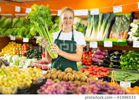 Portrait of a positive girl who works part-time as a trainee saleswoman, holding celery Portrait of a positive girl who works part-time as a trainee saleswoman, holding celery 91996533
