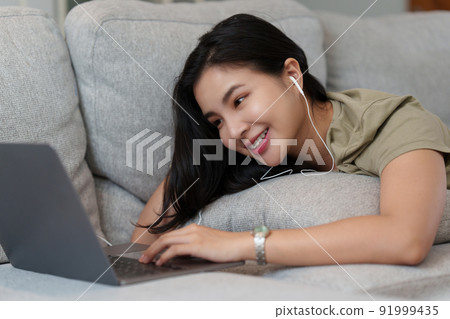 Portrait of Happy Beautiful woman lay down on cozy couch and listen music by streaming on laptop. 91999435