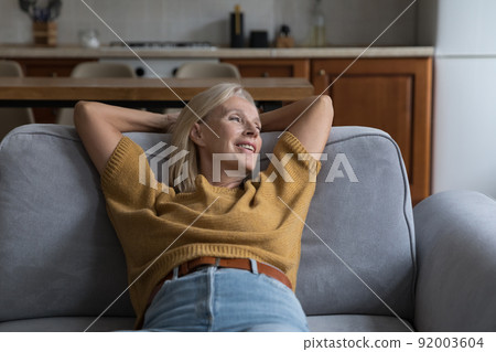 Smiling older female resting on sofa looks aside feels peaceful Smiling older female resting on sofa looks aside feels peaceful 92003604