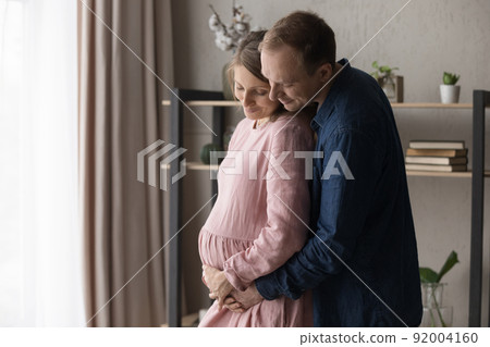 Loving future parents expecting their first baby hugging standing indoor 92004160
