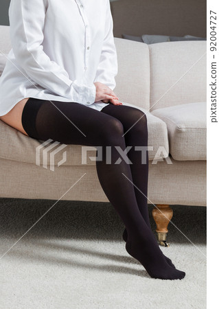 Black compression stockings on a woman in a white room. Black tights. Girl putting on stockings at home. Beautiful female legs. 92004727