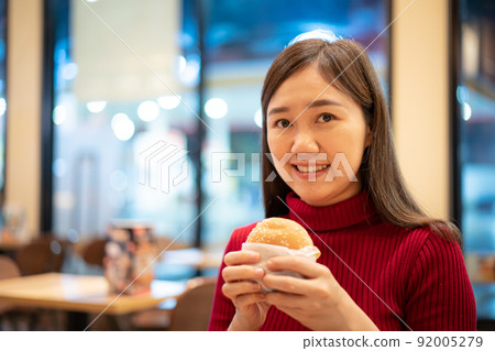 Cheerful Asian woman enjoy eating a grilled meat hamburger in the fast food restaurant at night. Unhealthy eating behaviour concept. Hungry woman holding a hamburger close up. Cheerful Asian woman enjoy eating a grilled meat hamburger in the fast food restaurant at night. Unhealthy eating behaviour concept. Hungry woman holding a hamburger close up. 92005279