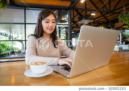 Asian beautiful working woman doing her work and drink in the coffee shop or cafe. Cheerful online blogger writing on her laptop or notebook computer and drinking a coffee close up. Modern lifestyle. Asian beautiful working woman doing her work and drink in the coffee shop or cafe. Cheerful online blogger writing on her laptop or notebook computer and drinking a coffee close up. Modern lifestyle. 92005283