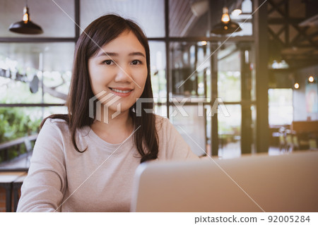 Asian beautiful working woman doing her work and drink in the coffee shop or cafe. Cheerful online blogger writing on her laptop or notebook computer and drinking a coffee close up. Modern lifestyle. Asian beautiful working woman doing her work and drink in the coffee shop or cafe. Cheerful online blogger writing on her laptop or notebook computer and drinking a coffee close up. Modern lifestyle. 92005284