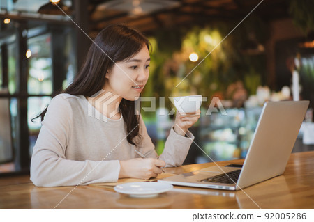 Asian beautiful working woman doing her work and drink in the coffee shop or cafe. Cheerful online blogger writing on her laptop or notebook computer and drinking a coffee close up. Modern lifestyle. Asian beautiful working woman doing her work and drink in the coffee shop or cafe. Cheerful online blogger writing on her laptop or notebook computer and drinking a coffee close up. Modern lifestyle. 92005286