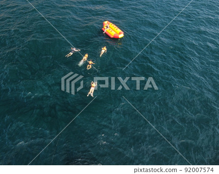 Happy people swim on air mattress behind a high speed water bike. Tourists ride on inflatable watercraft boat. Sea attraction. Water scooter skating people on large Inflatable raft. Aerial copter view 92007574
