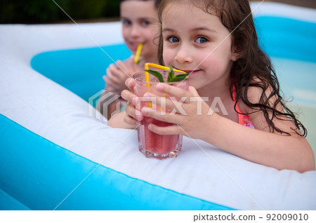 Charming Caucasian little girl drinks fresh cocktail and smiles looking at camera while having fun in an inflatable pool Charming Caucasian little girl drinks fresh cocktail and smiles looking at camera while having fun in an inflatable pool 92009010
