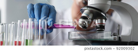 Female scientist pouring pink sample liquid on glass container under microscope in lab 92009302