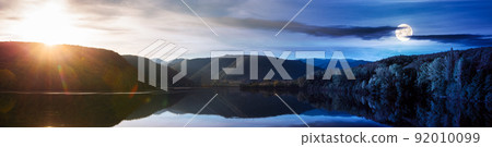 day and night time change above beautiful autumn landscape by the lake. trees on the shore and forested mountains reflecting in the water surface. sun and moon above horizon 92010099