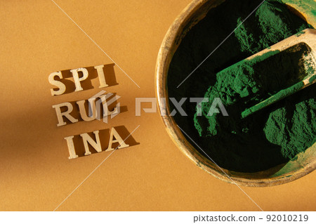 Blue-green algae spirulina powder in bamboo eco bowl with text SPIRULINA. Super powder in spoon. Food supplement source of protein and beta carotene 92010219