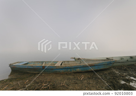 Two old blue weathered shabby retro boats on the riverbank in thick fog. Cottagecore or unplugged concept. Countryside calm soothing landscape. Copy space. Horizontal orientation. Two old blue weathered shabby retro boats on the riverbank in thick fog. Cottagecore or unplugged concept. Countryside calm soothing landscape. Copy space. Horizontal orientation. 92010803