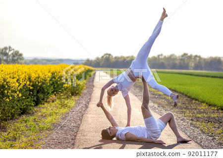 Beautiful young couple doing acro yoga on the bike path. Man lying on road and balancing woman in his feet. Beautiful young couple doing acro yoga on the bike path. Man lying on road and balancing woman in his feet. 92011177