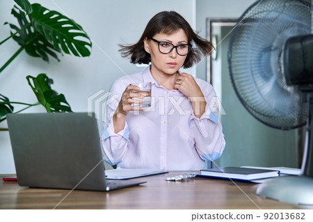 Middle-aged woman suffering from heat, overheated, cooled by fan and fan 92013682