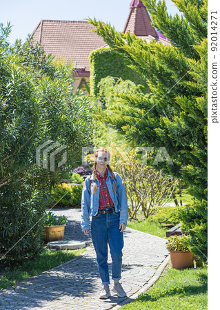 A smiling woman in a denim outfit walks along a paved park path passing between lawns planted with dense trees and bushes on a sunny spring day A smiling woman in a denim outfit walks along a paved park path passing between lawns planted with dense trees and bushes on a sunny spring day 92014271