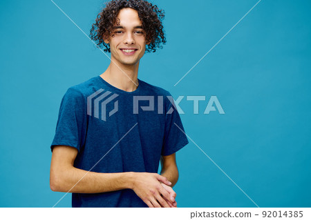 Smiling joyful cute curly tanned Caucasian guy in basic t-shirt posing isolated on over blue background. Lifestyle and Emotions concept. Good offer with copy space for ad 92014385