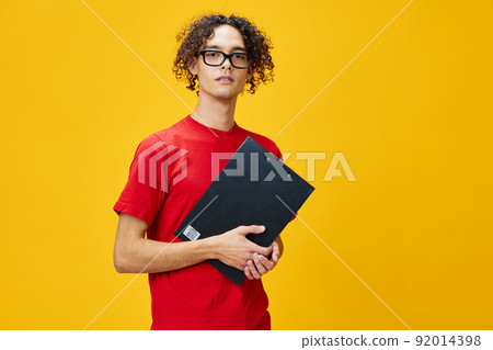 Confused myopic young student man in red t-shirt funny eyewear holds tablet folder with study notes posing isolated on over yellow studio background. Free place for ad. Education College concept Confused myopic young student man in red t-shirt funny eyewear holds tablet folder with study notes posing isolated on over yellow studio background. Free place for ad. Education College concept 92014398
