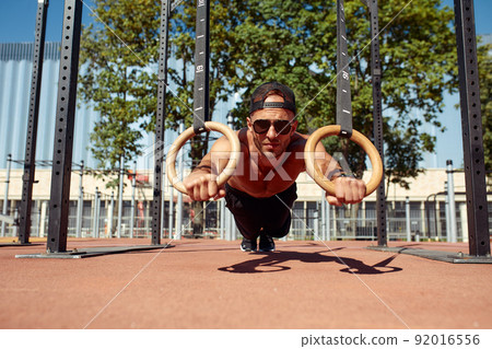 Muscular sportsman exercising on outdoor gymnastic rings in outdoor gym. Hands at rings dipping man doing exercise using rings 92016556