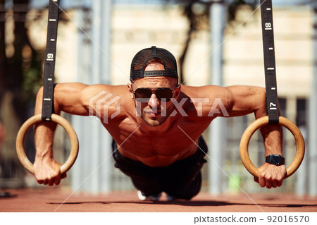 Muscular sportsman exercising on outdoor gymnastic rings in outdoor gym. Hands at rings dipping man doing exercise using rings 92016570