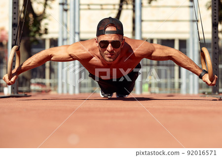 Handsome man on sport ground. The sportsman the guy, carries out difficult exercise, sports gymnastics. Muscular man doing workout with rings. The concept of a modern young man Handsome man on sport ground. The sportsman the guy, carries out difficult exercise, sports gymnastics. Muscular man doing workout with rings. The concept of a modern young man 92016571