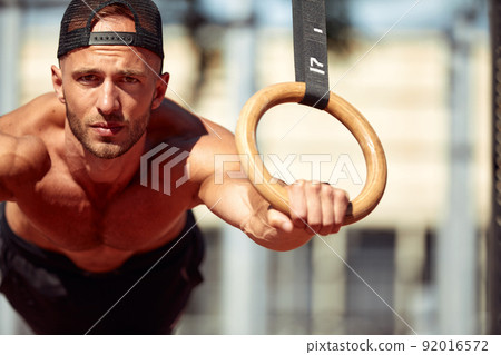 Muscular sportsman exercising on outdoor gymnastic rings in outdoor gym. Hands at rings dipping man doing exercise using rings Muscular sportsman exercising on outdoor gymnastic rings in outdoor gym. Hands at rings dipping man doing exercise using rings 92016572