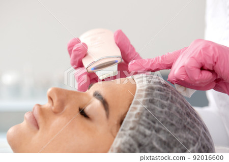 Cosmetologist, beautician in pink gloves making facial treatment with ultrasonic spatula to woman, face skin scrubber treatment with ultrasonic spatula, facial cleansing procedure in beauty salon 92016600