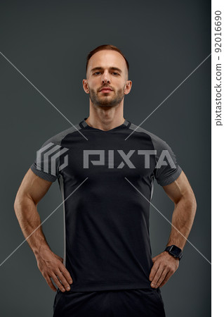 Half length portrait of young sporty guy in t-shirt and shorts standing on gray studio background. Serious millennial sportsman looking at camera. Healthy lifestyle and sports concept 92016690