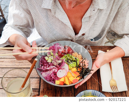 Woman eating tasty colorful healthy natural organic vegetarian Hawaiian poke bowl using asian chopsticks on rustic wooden table. Healthy natural organic eating concept Woman eating tasty colorful healthy natural organic vegetarian Hawaiian poke bowl using asian chopsticks on rustic wooden table. Healthy natural organic eating concept 92017221