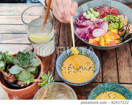 Woman eating tasty colorful healthy natural organic vegetarian Hawaiian poke bowl using asian chopsticks on rustic wooden table. Healthy natural organic eating concept Woman eating tasty colorful healthy natural organic vegetarian Hawaiian poke bowl using asian chopsticks on rustic wooden table. Healthy natural organic eating concept 92017222