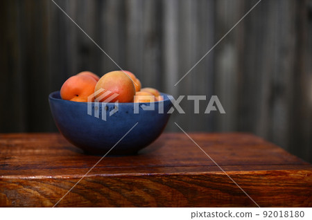 Ripe apricot fruits in a blue ceramic bowl standing on a wooden surface against wood background Ripe apricot fruits in a blue ceramic bowl standing on a wooden surface against wood background 92018180