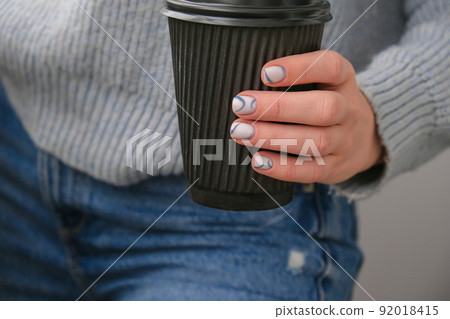 Manicured female hands holding black eco recycling coffee cup with stylish blue nails and minimalistic design. Trendy modern design manicure. Gel nails. Skin care. Beauty treatment. Nail care 92018415