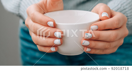 Manicured female hands holding white coffee cup with stylish blue nails and minimalistic design. Trendy modern design manicure. Gel nails. Skin care. Beauty treatment. Nail care Beautician 92018718
