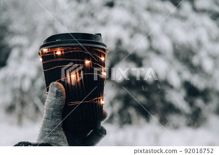 Female hand in winter warm gloves holding black eco paper cup with garland shiny light. Creative trendy zero waste recycle cup. Mockup. Hot warming drink to go in winter day. Take away concept 92018752